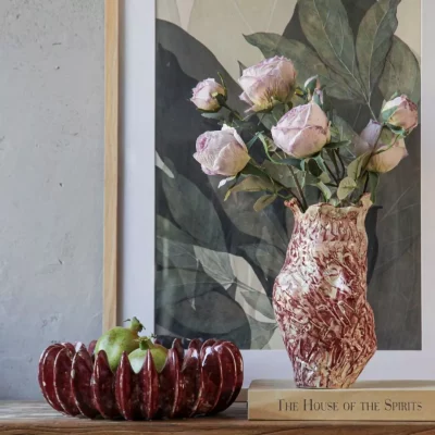 Artisanal marbled red-and-white ceramic vase with a textured, organic shape, holding a bouquet of dried pink roses, styled on a wooden book titled 