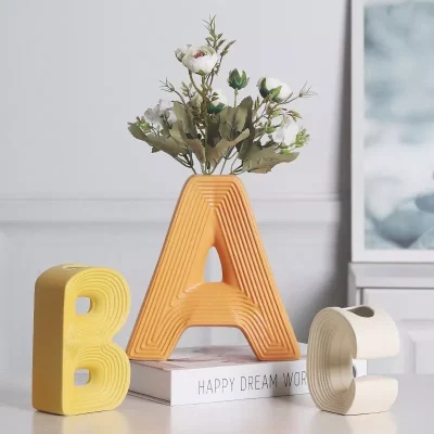 Vibrant A, B, C letter-shaped ceramic vases in yellow, orange, and cream, with the 'A' vase holding a bouquet of artificial white flowers, styled on a modern home desk.