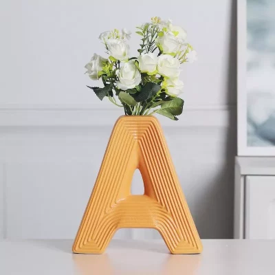 Vibrant A, B, C letter-shaped ceramic vases in yellow, orange, and cream, with the 'A' vase holding a bouquet of artificial white flowers, styled on a modern home desk.