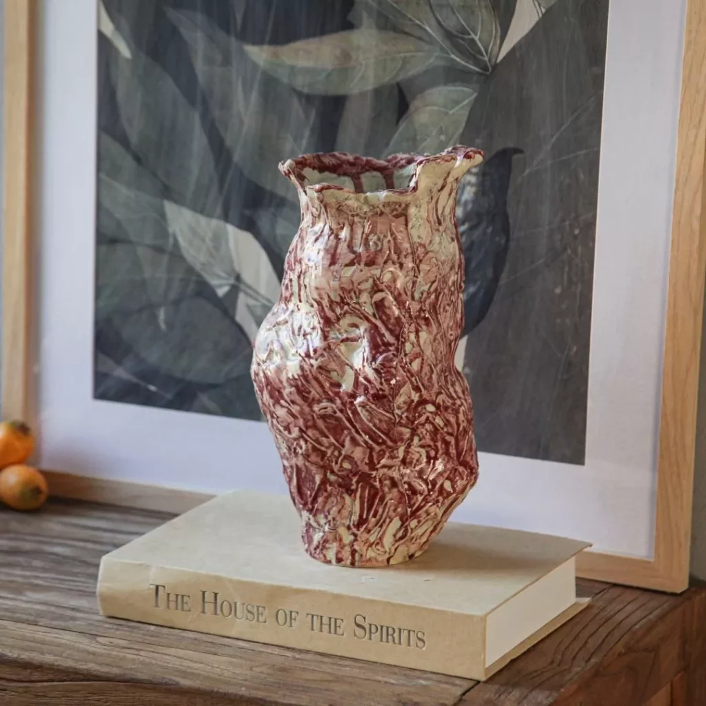 Artisanal marbled red-and-white ceramic vase with a textured, organic shape, holding a bouquet of dried pink roses, styled on a wooden book titled 