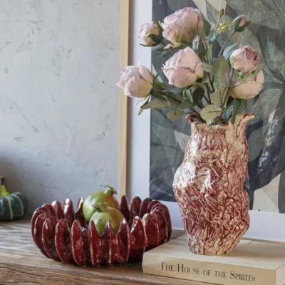 Artisanal marbled red-and-white ceramic vase with a textured, organic shape, holding a bouquet of dried pink roses, styled on a wooden book titled 