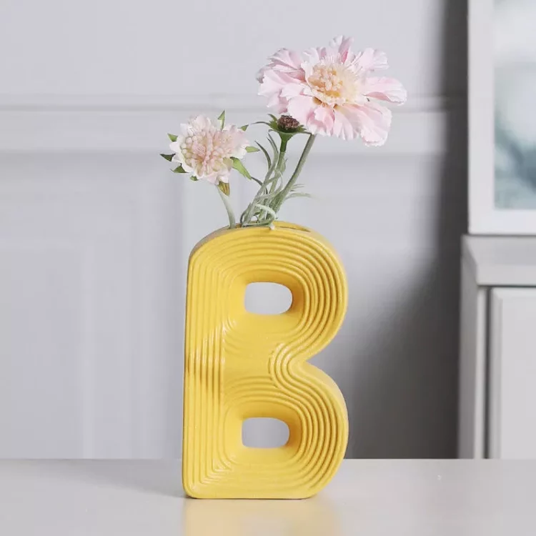 Vibrant A, B, C letter-shaped ceramic vases in yellow, orange, and cream, with the 'A' vase holding a bouquet of artificial white flowers, styled on a modern home desk.