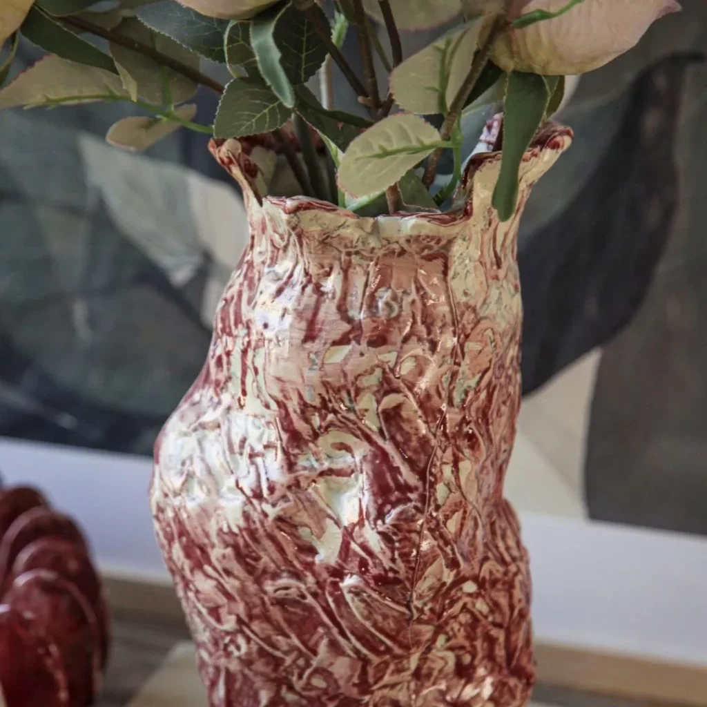 Artisanal marbled red-and-white ceramic vase with a textured, organic shape, holding a bouquet of dried pink roses, styled on a wooden book titled 