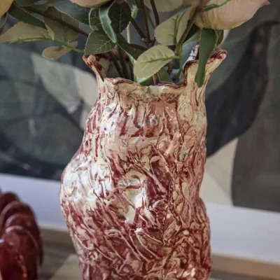 Artisanal marbled red-and-white ceramic vase with a textured, organic shape, holding a bouquet of dried pink roses, styled on a wooden book titled 